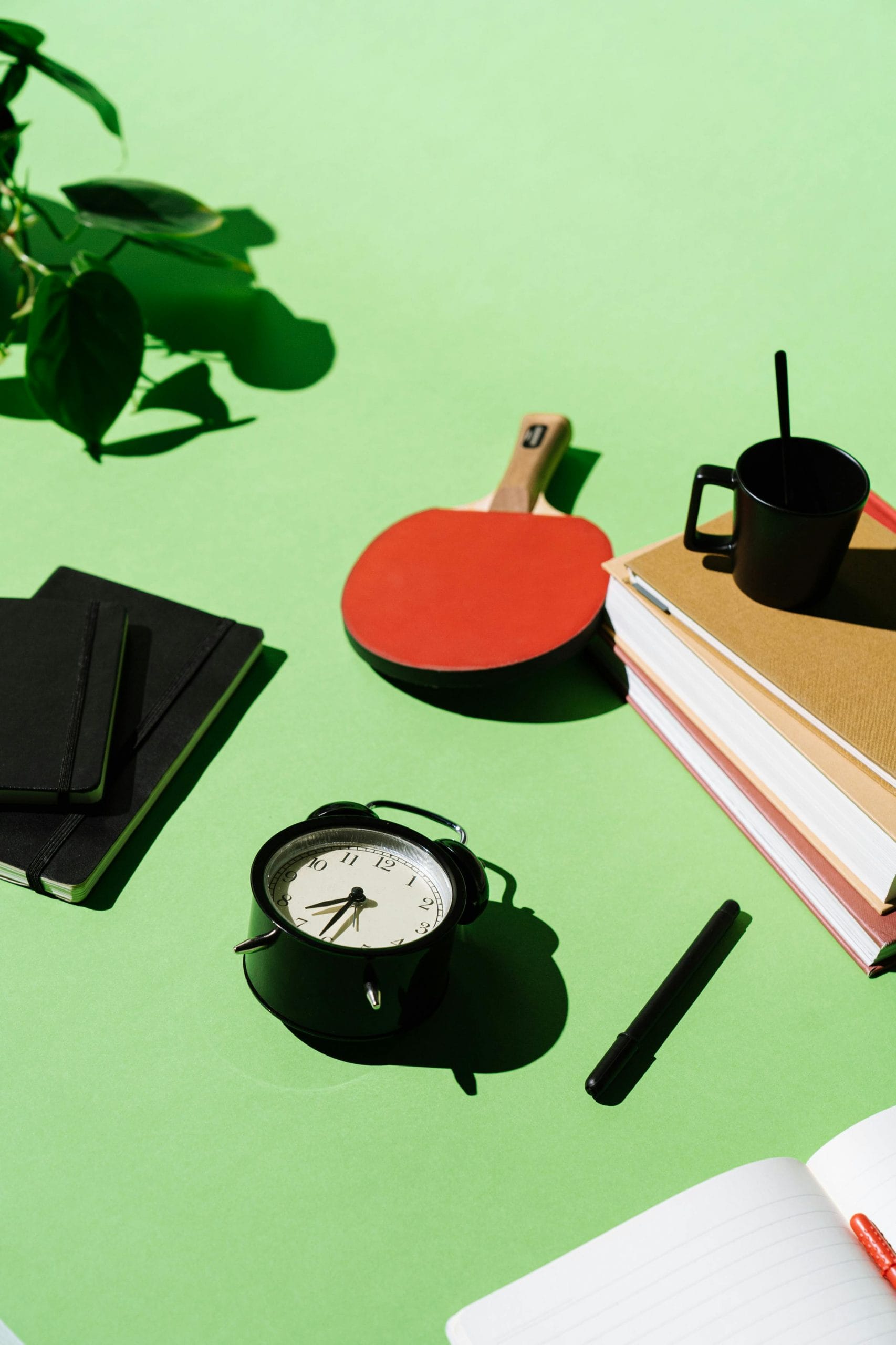 A minimal workspace with books, alarm clock, racket, and greenery on a vibrant green background.
